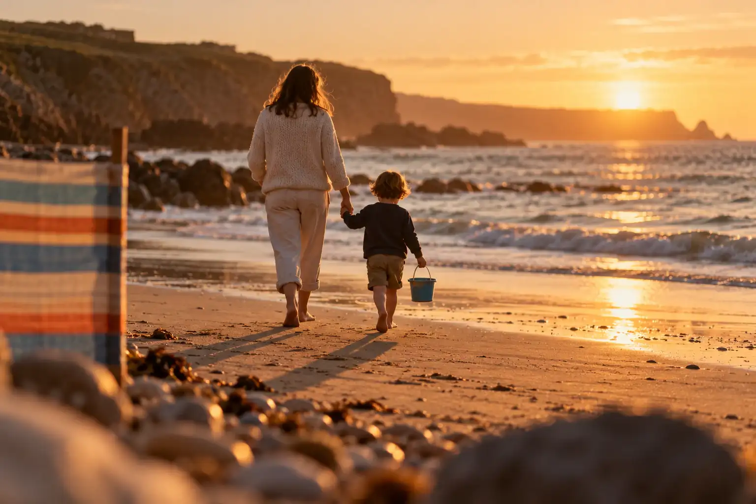 A parent and a small child seen from behind, walking hand in hand along a beach at sunset.